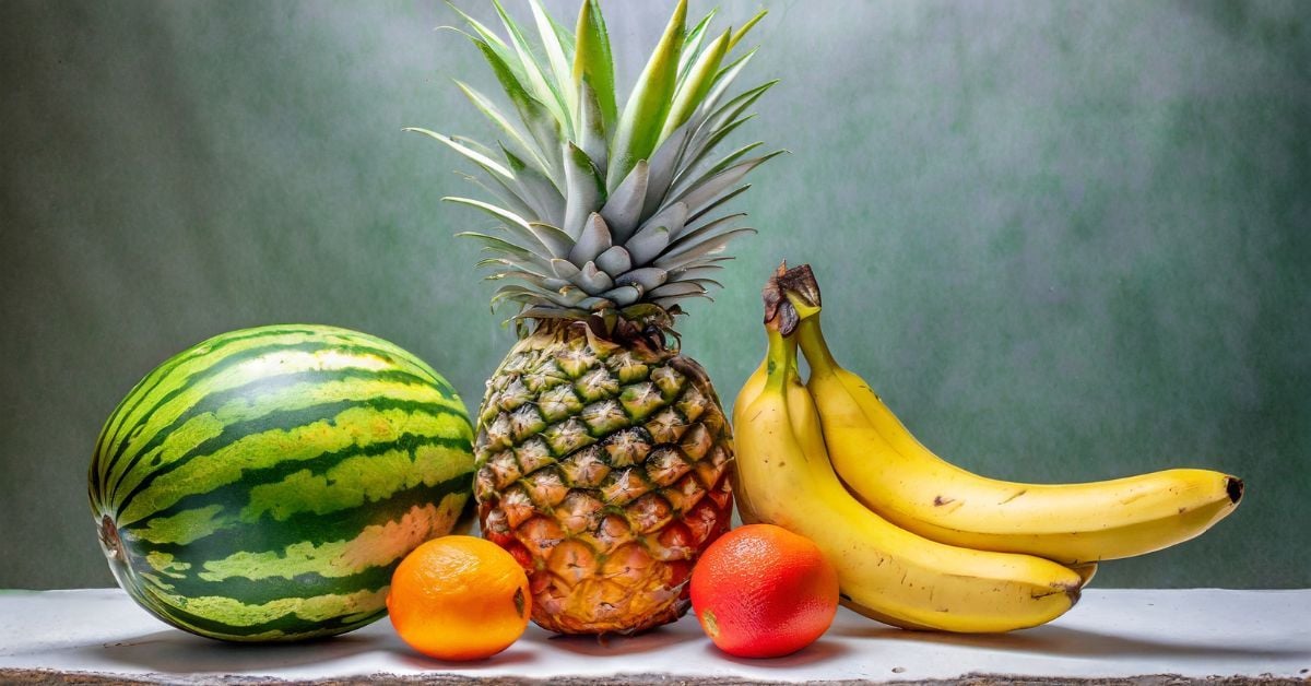 A vibrant selection of fruits is arranged against a green background. The display features a watermelon, two oranges, a pineapple, and a bunch of bananas, creating a colorful and appetizing composition that celebrates freshness and variety.